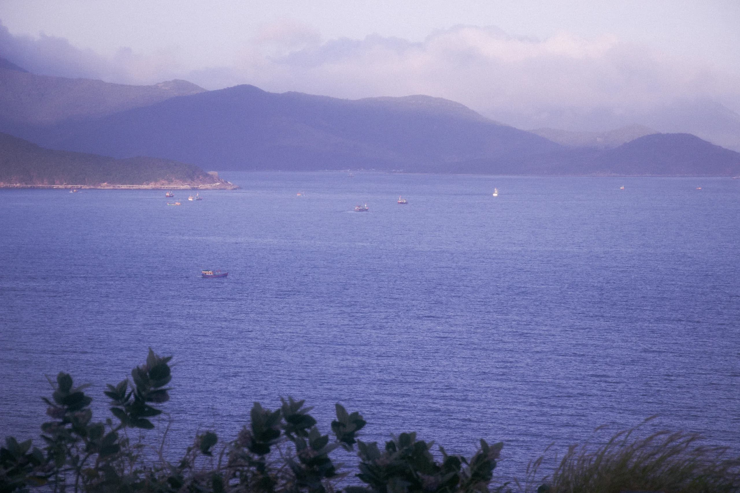 Mist settling over a calm sapphire-blue cove, with small fishing boats resting on still water beneath distant mountains.